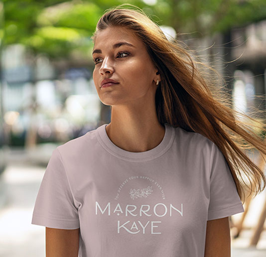 A young woman wearing a light-colored t-shirt with the Marron Kaye logo, walking outdoors with a confident expression.
