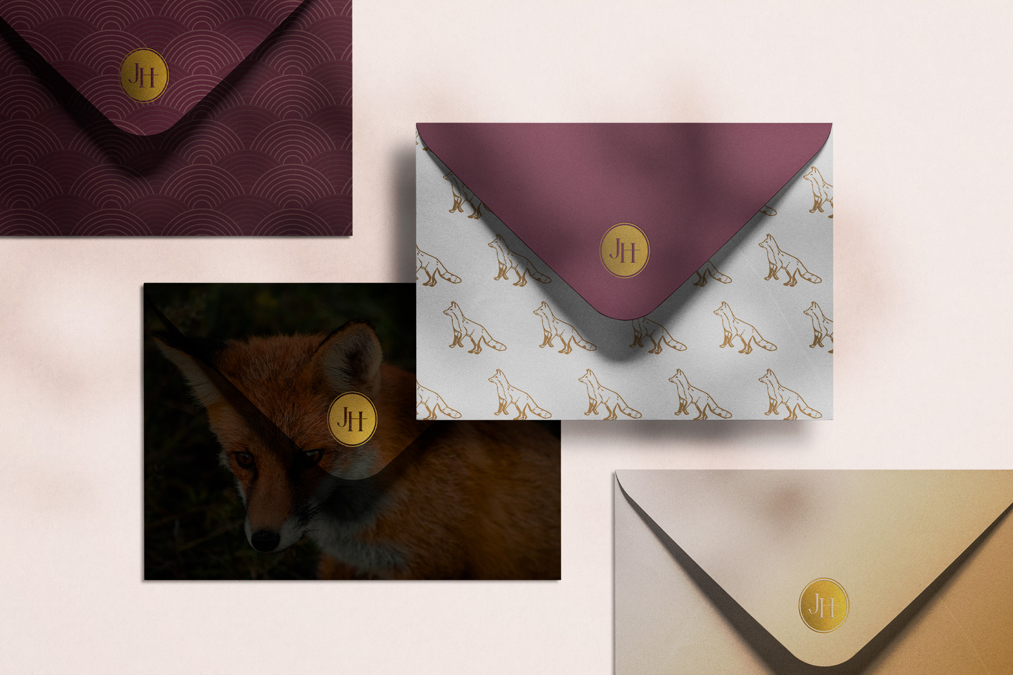 A collection of envelopes with sophisticated designs for Juniper Hartmann. The envelopes feature patterns such as golden fox illustrations, deep burgundy scallop patterns, and a photograph of a fox. Each envelope is sealed with a gold foil sticker featuring the JH logo.