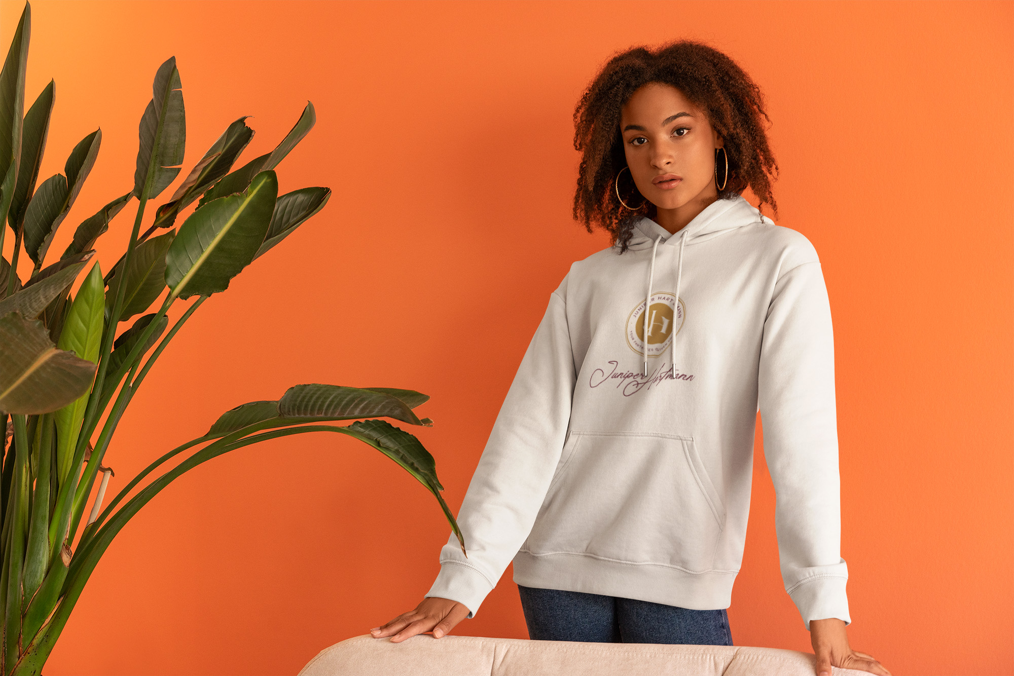 A stylish young woman wearing a white hoodie featuring the Juniper Hartmann logo and signature. The background is a vibrant orange, with green houseplants adding a modern and warm touch.