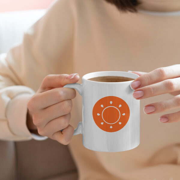 A person holding a white ceramic mug featuring a bold orange sun logo.